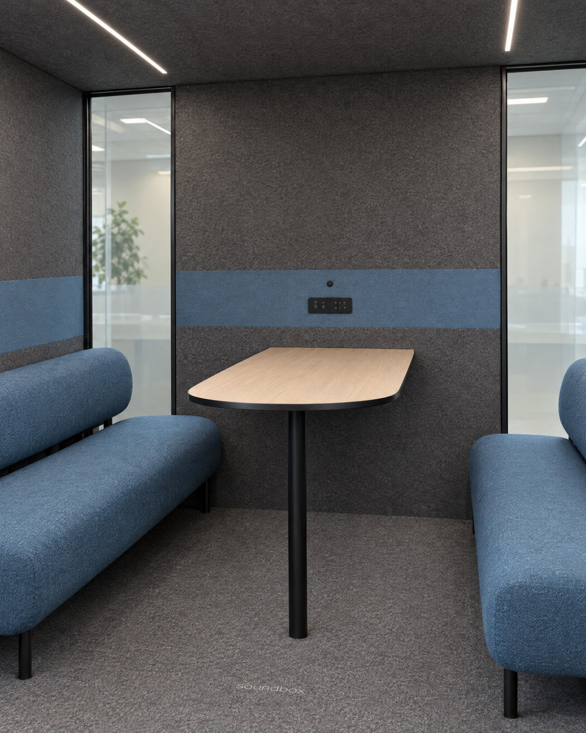 Inside the SoundBox VRS Collaborative from the open door: a wide room with two dusty blue sofas facing each other, an oak table from the middle of the back wall, control panel on the felt, glass at both ends of the back wall. Illustration.