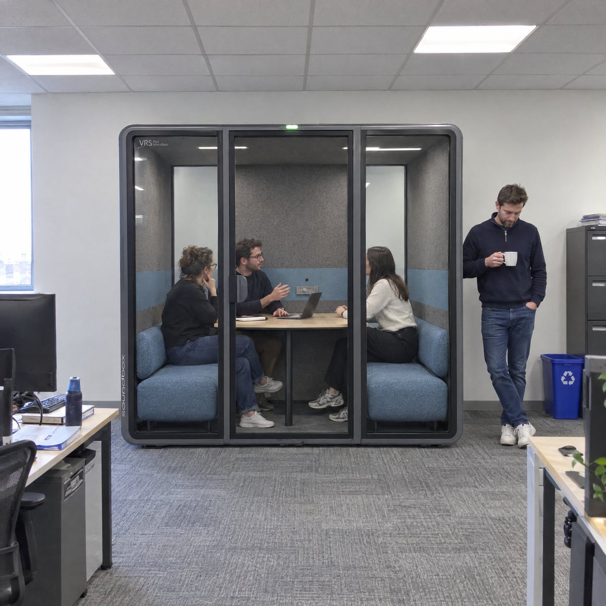 Soundbox VRS Collaborative against an office wall with three people meeting inside and a colleague standing beside it