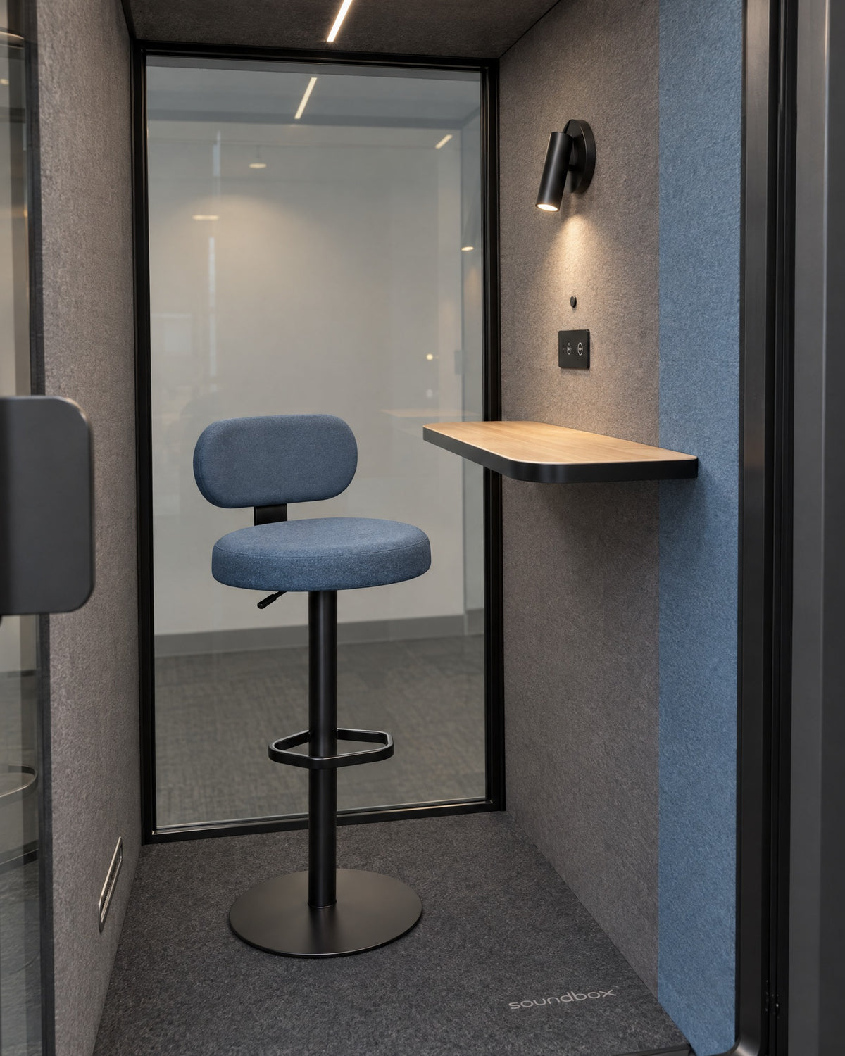 Inside the SoundBox VRS Single through the open door: dusty blue bar stool, oak desk shelf, spot lamp and control panel, full-height glass back wall. Illustration.