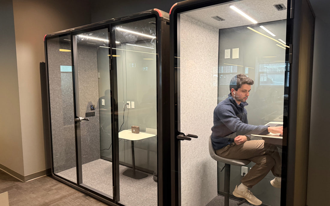 A man wearing headphones working inside a Quiet Cabins room on an office floor, door open