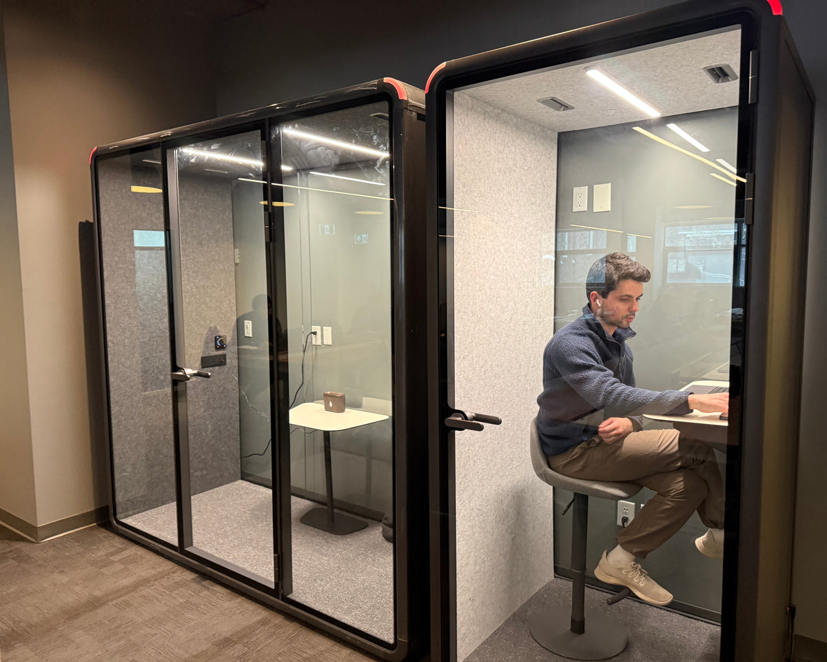 A man wearing headphones working inside a Quiet Cabins room on an office floor, door open