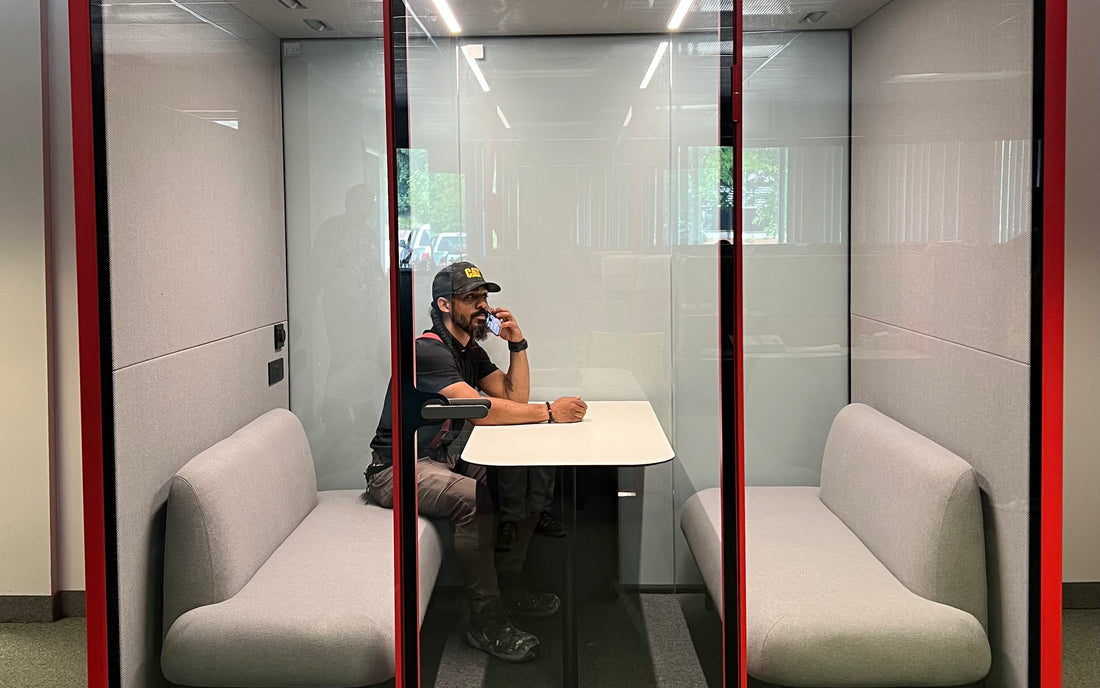 A man on a phone call inside a closed Quiet Cabins booth on an office floor
