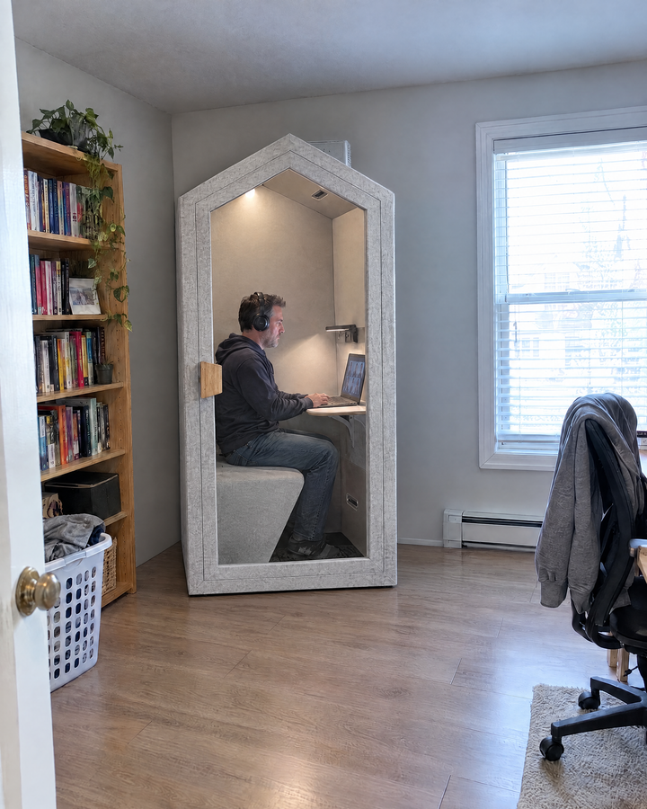Home Pod pushed against the wall of a spare bedroom office, a man on a video call inside with the door closed. Illustration.