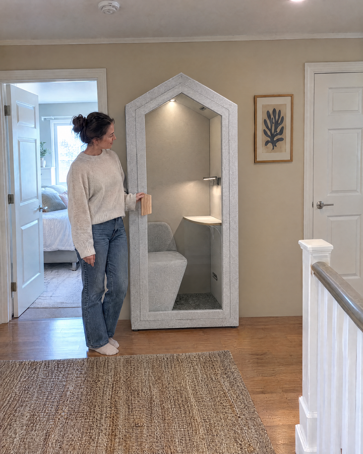 A woman standing beside the Home Pod, which stands against the wall of an upstairs landing next to a bedroom door for scale. Illustration.