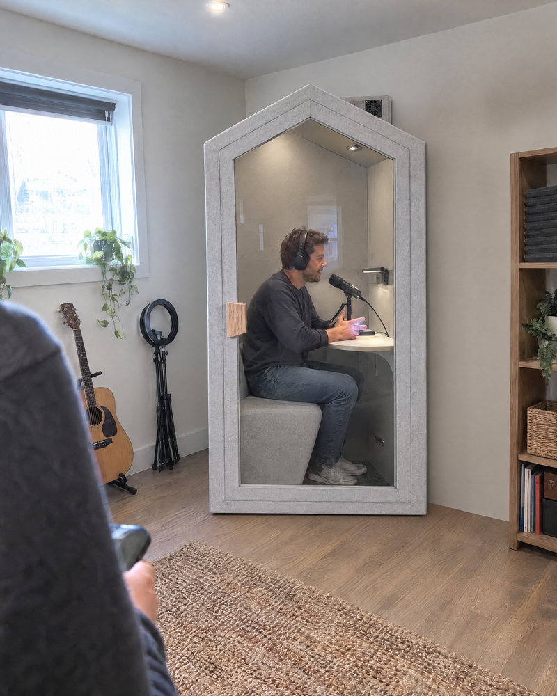 A man recording a podcast into a desk microphone inside the Home Pod, set against the wall of a spare room. Illustration.