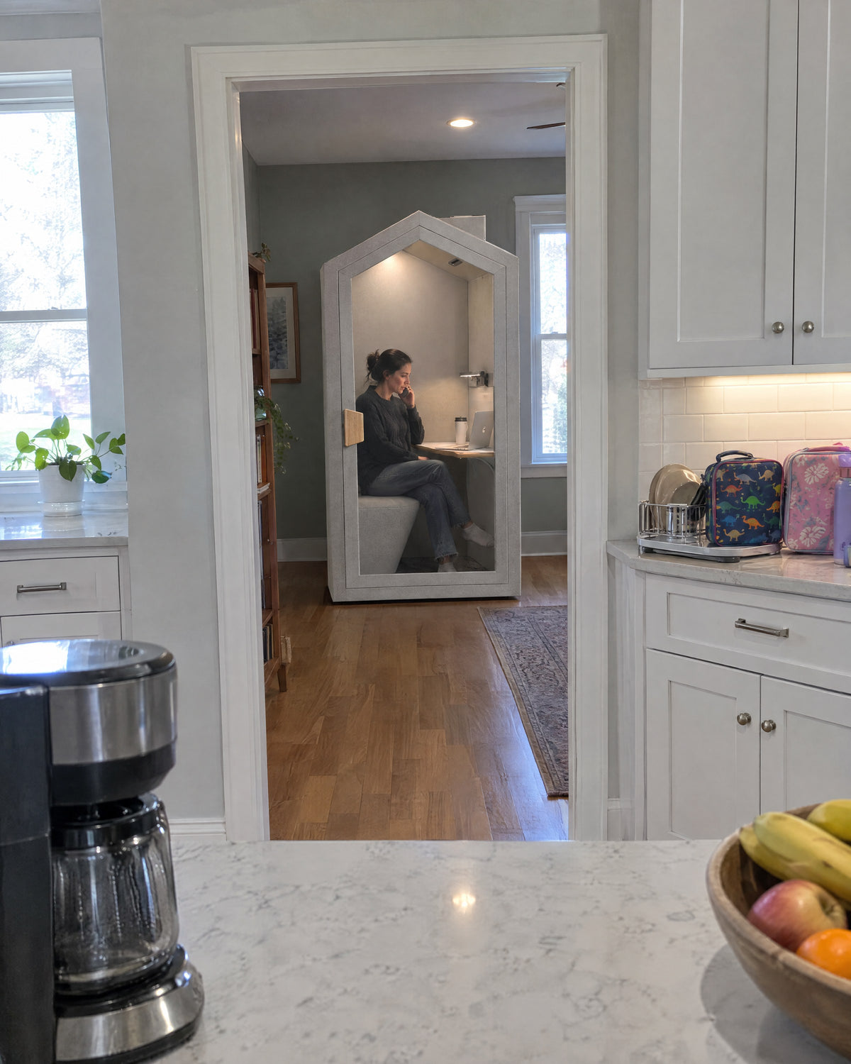 Home Pod in a den seen through the kitchen doorway, a woman on a call inside. Illustration.