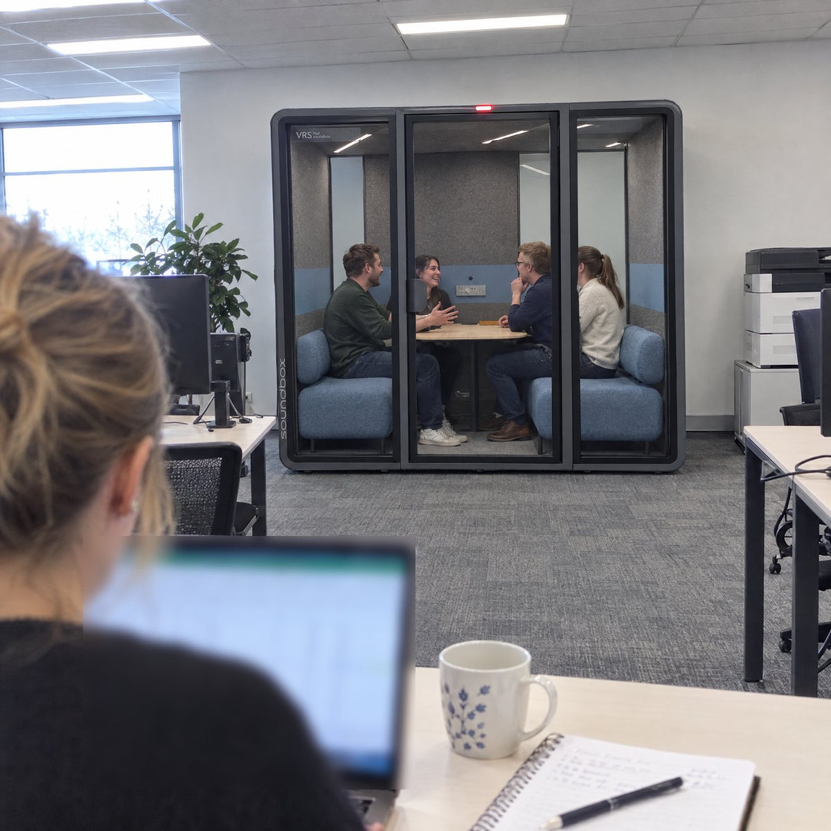 A colleague works at her desk while four people talk inside a Soundbox VRS Collaborative behind her, door closed