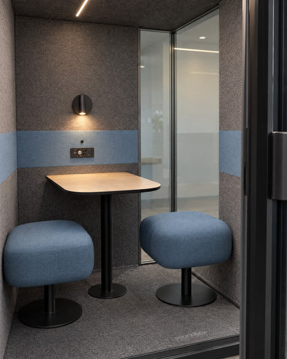 Inside the SoundBox VRS Comfort through the open door: two backless dusty blue seats and a small oak table, wall lamp and control panel on the felt back wall, glass on the right half of the back wall. Illustration.