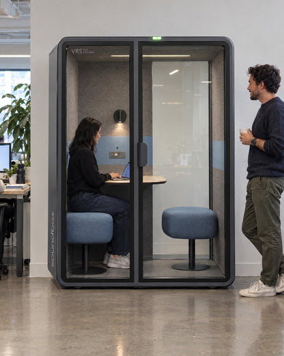 SoundBox VRS Comfort in an open-plan office, one person inside on a video call, a colleague standing outside. Illustration.