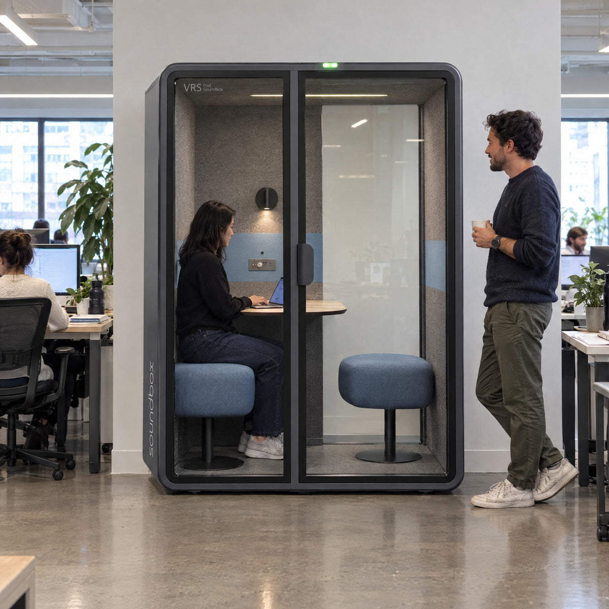 SoundBox VRS Comfort in an open-plan office, one person inside on a video call, a colleague standing outside. Illustration.