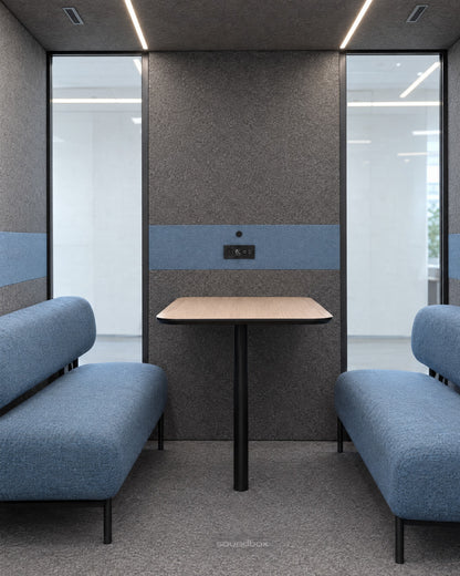 Inside the SoundBox VRS Double from the open door: a wide, shallow room with two dusty blue sofas facing each other, an oak table at the middle of the back wall, control panel on the felt, wide glass at both ends of the back wall. Illustration.