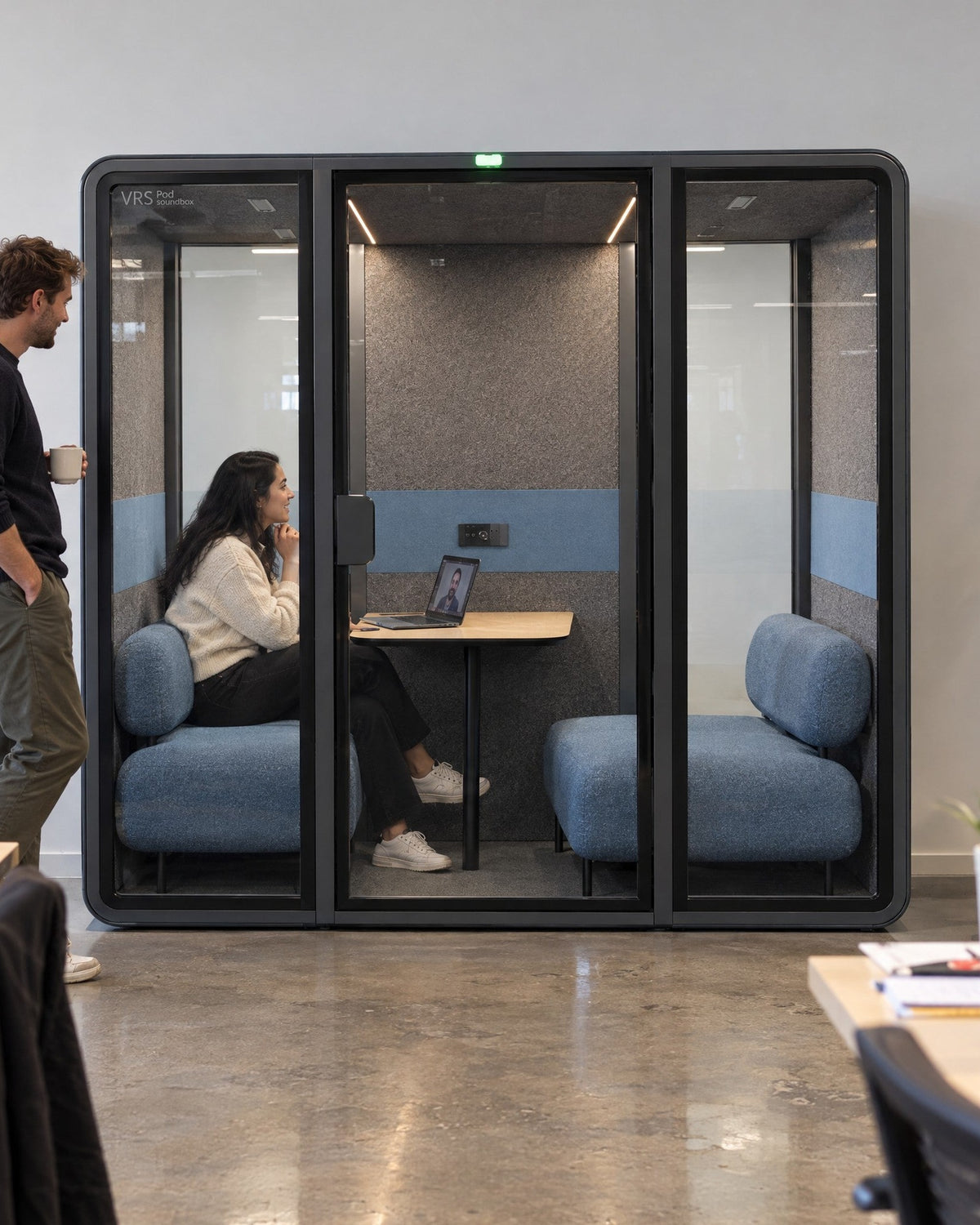 SoundBox VRS Double against the wall of an open-plan office, one person inside on a video call, a colleague standing outside. Illustration.