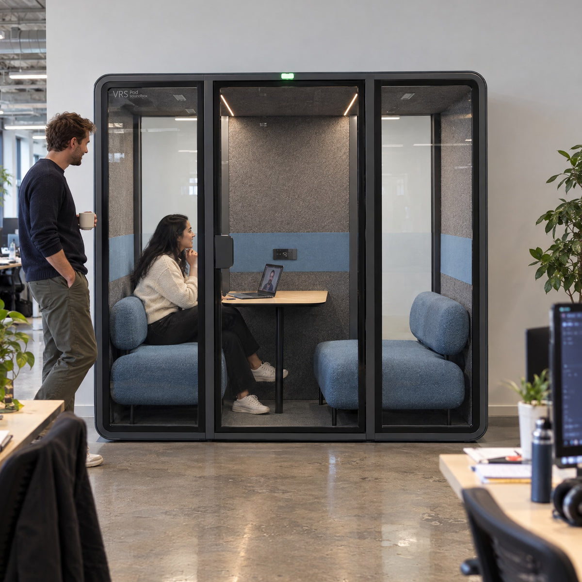 SoundBox VRS Double against the wall of an open-plan office, one person inside on a video call, a colleague standing outside. Illustration.