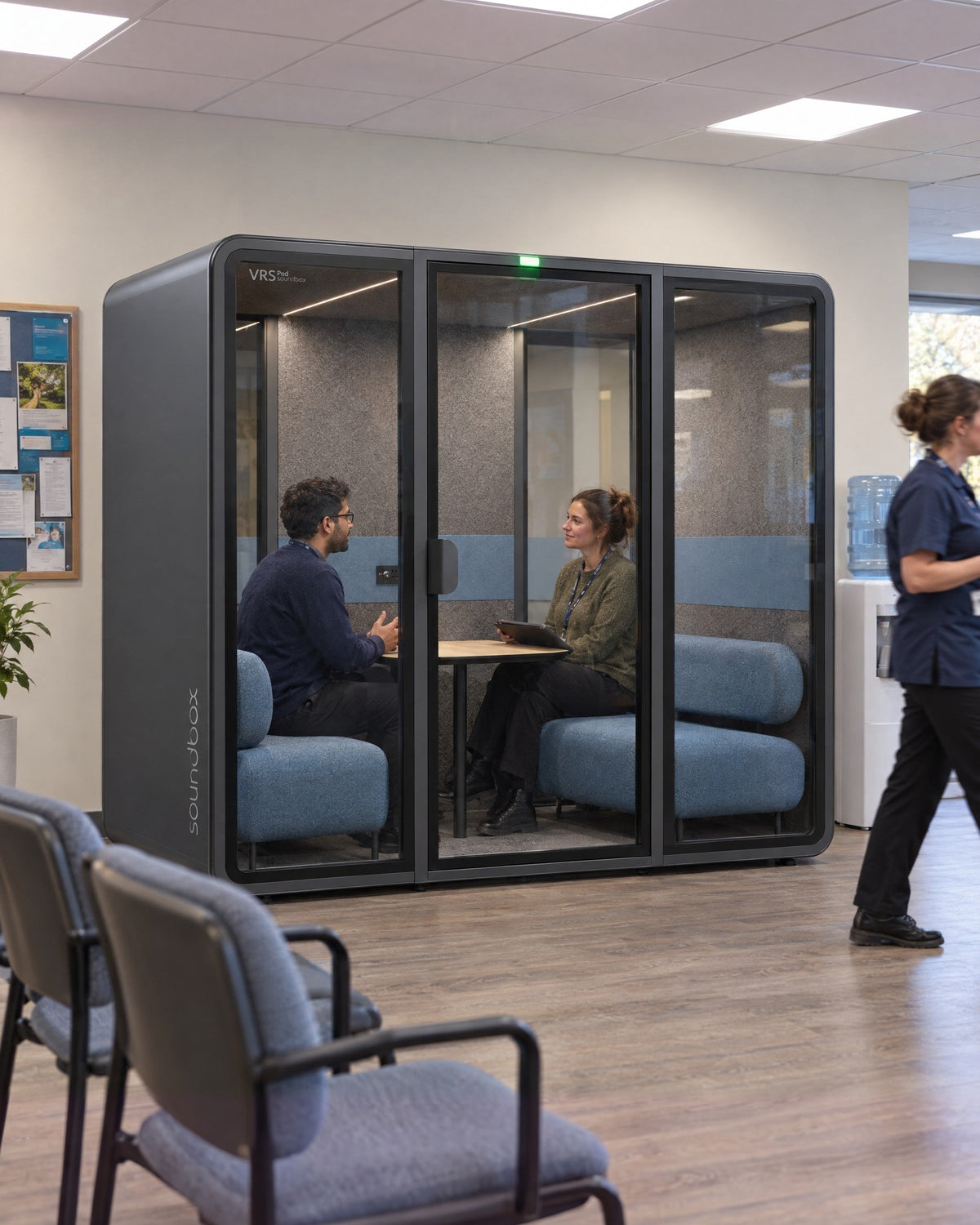 SoundBox VRS Double in a community clinic staff area seen from the waiting chairs, two people in a private conversation inside. Illustration.