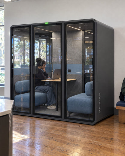 SoundBox VRS Double in a sunlit university staff area, one person inside on a video call with headphones, a student on a bench nearby. Illustration.