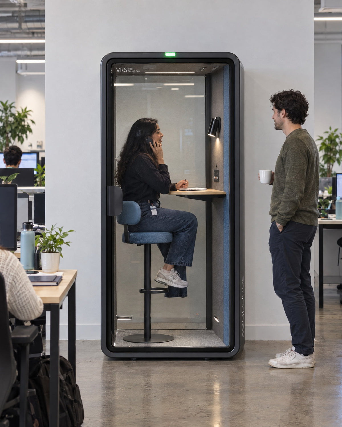 SoundBox VRS Single in an open-plan office, one person inside on a phone call on the bar stool, a colleague outside. Illustration.