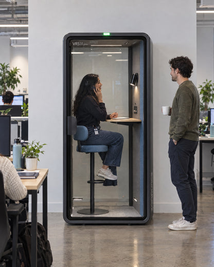 SoundBox VRS Single in an open-plan office, one person inside on a phone call on the bar stool, a colleague outside. Illustration.