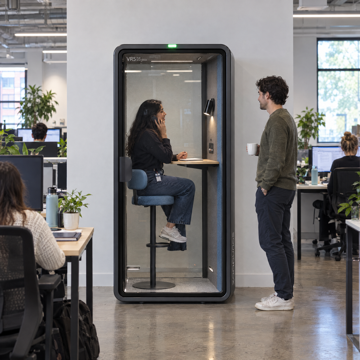 SoundBox VRS Single in an open-plan office, one person inside on a phone call on the bar stool, a colleague outside. Illustration.