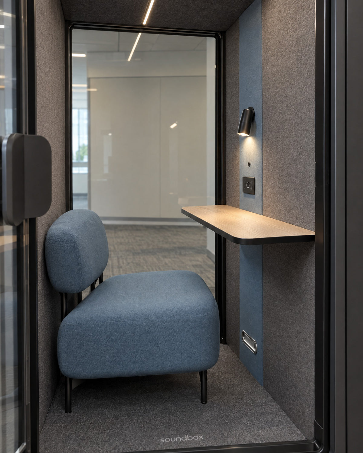 Inside the SoundBox VRS Single Comfort through the open door: dusty blue lounge armchair, oak desk shelf, spot lamp and control panel, full-height glass back wall. Illustration.