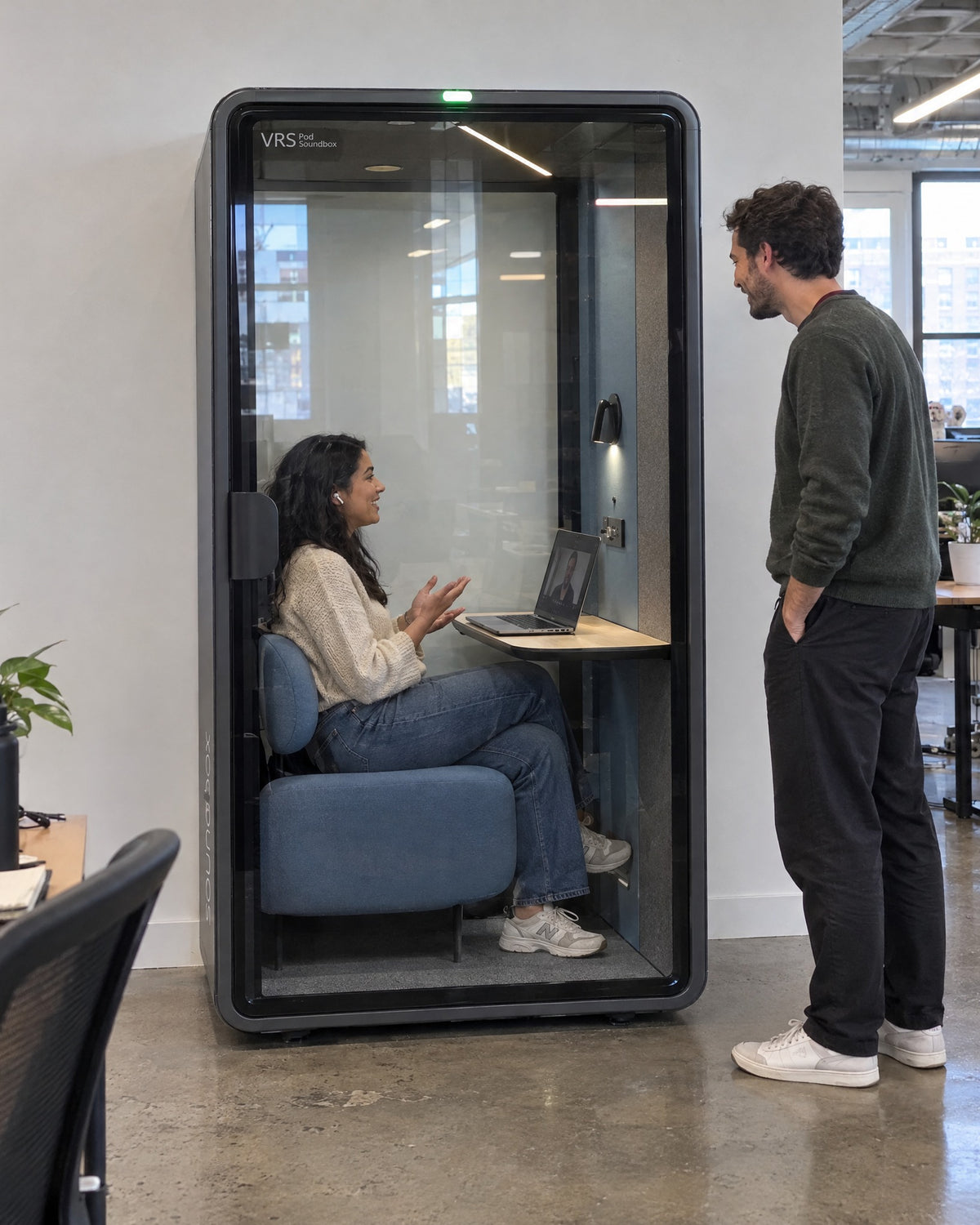 SoundBox VRS Single Comfort in an open-plan office, one person inside on a video call, a colleague standing outside. Illustration.