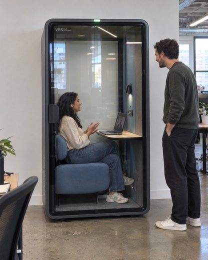 SoundBox VRS Single Comfort in an open-plan office, one person inside on a video call, a colleague standing outside. Illustration.