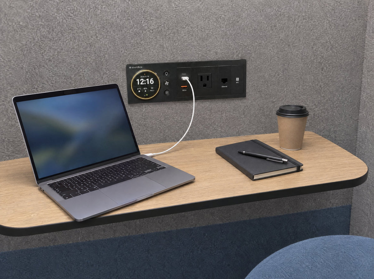 Light oak desk shelf inside a SoundBox VRS pod with a laptop, notebook and coffee, the smart panel with a standard outlet above it